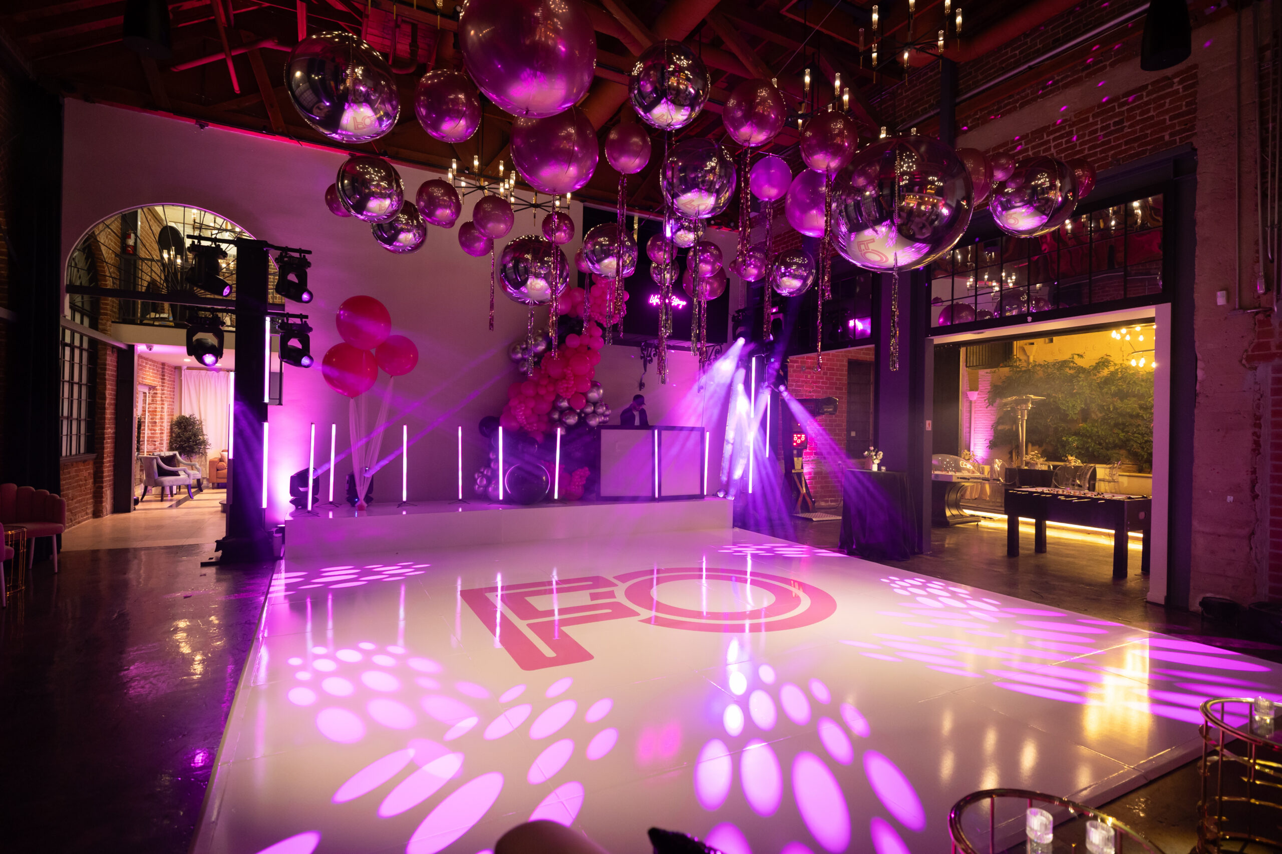 8JanBirch979A5877 - Bronson Hospitality Indoor party venue with pink/purple lighting, oversized chrome balloons, and a glossy dance floor with a DJ booth in the background.