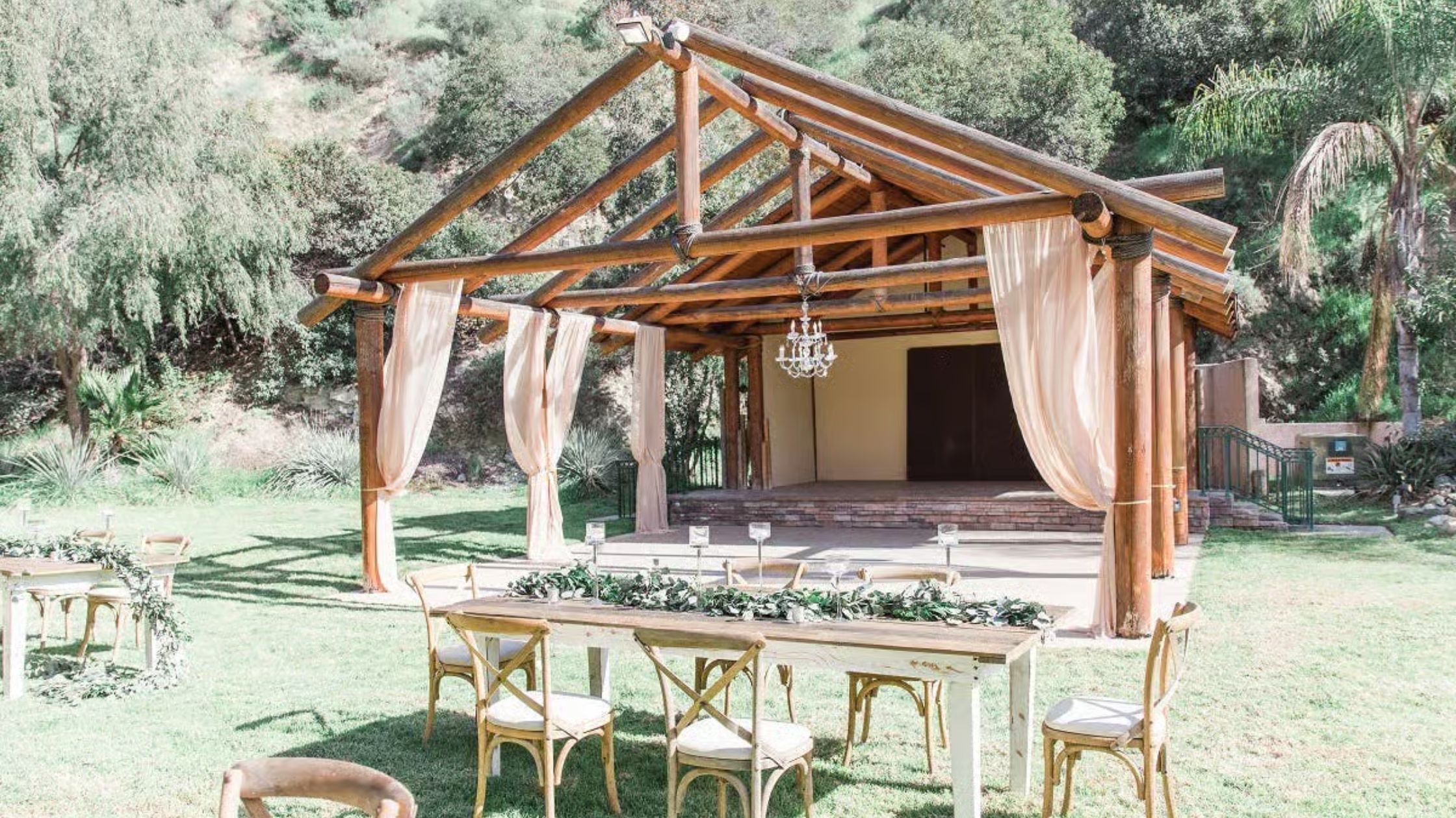 Castaway Burbank - Bronson Hospitality A charming outdoor garden space at Castaway Garden in Burbank featuring a rustic wooden pavilion with flowing drapery, elegant seating arrangements, and lush greenery surrounded by scenic natural landscapes