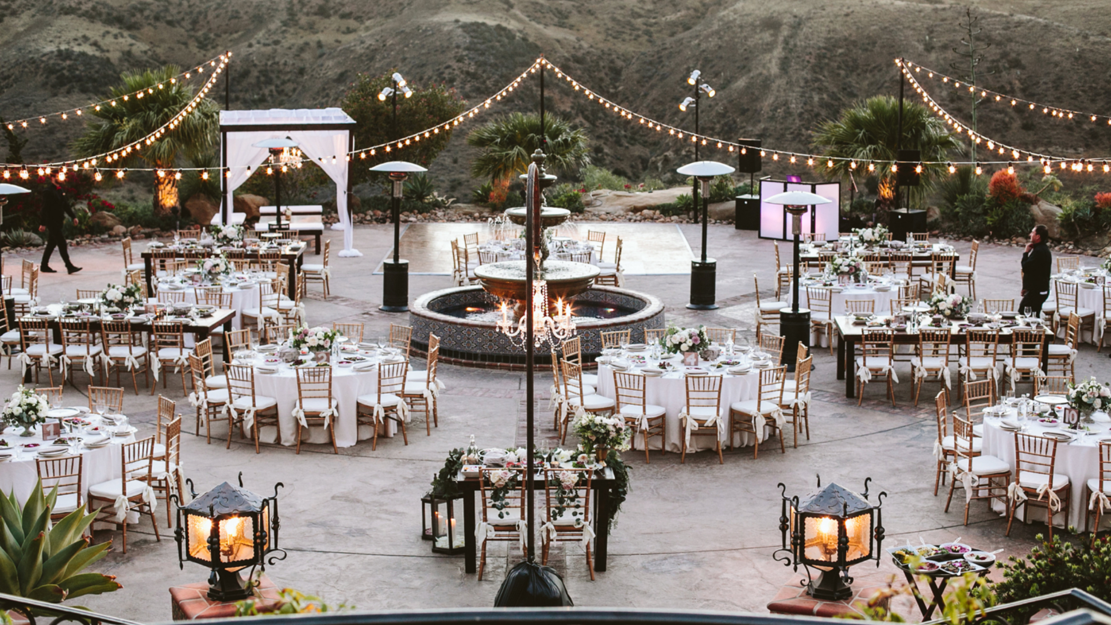 Hummingbird Nest Ranch - Bronson Hospitality Outdoor evening wedding reception with string lights, round and rectangular tables, and a central fountain backdrop set against a rocky hillside.