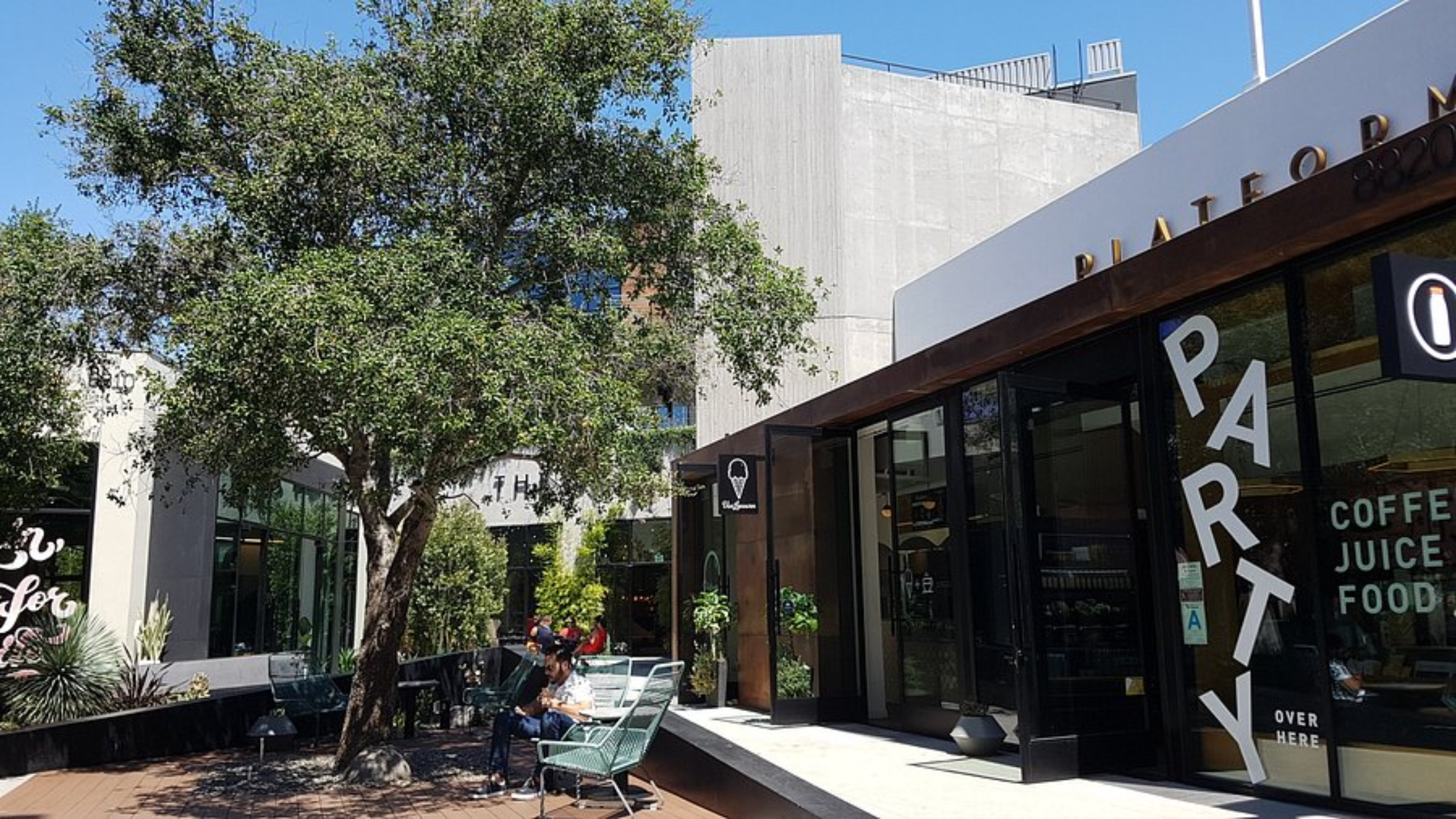 Platform LA - Bronson Hospitality Outdoor cafe area beside a modern concrete building with large trees and glass storefronts; people seated at tables under a clear blue sky.