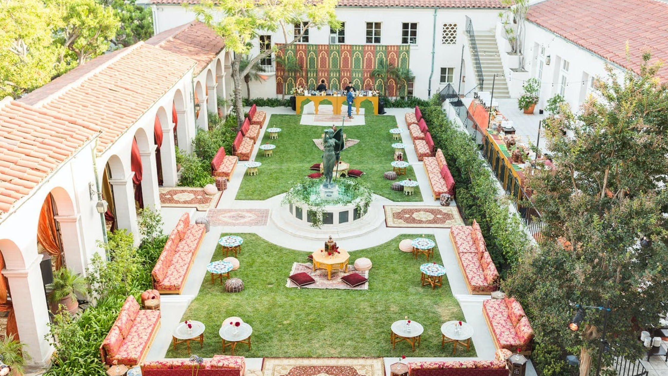 The Ebell of Los Angeles - Bronson Hospitality An elegant courtyard at The Ebell of Los Angeles featuring a central fountain, lush greenery, Mediterranean-style architecture, and colorful lounge seating arranged in a refined outdoor setting.