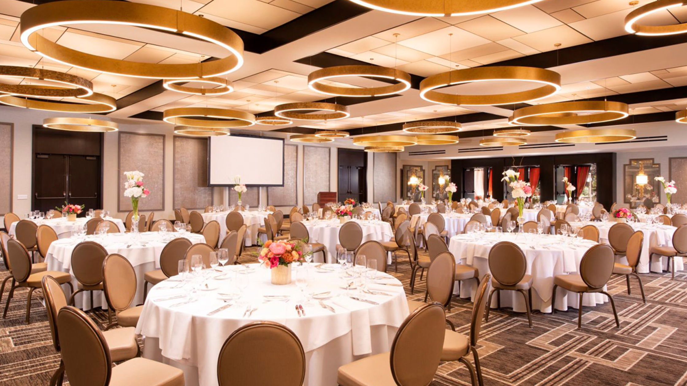 The Garland - Bronson Hospitality A modern event space at The Garland featuring round tables with white linens, floral centerpieces, contemporary circular lighting fixtures, and an elegant indoor reception setup