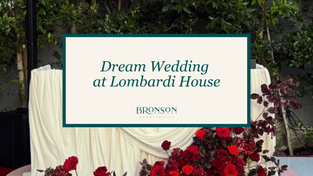 Beige wedding-sign reading 'Dream Wedding at Lombardi House' framed by dark teal border against a greenery backdrop with red flowers at the bottom.