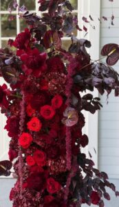 Screenshot - Bronson Hospitality Bouquet of deep burgundy flowers with red roses, dark anthuriums, and trailing burgundy leaves against a pale background.