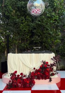 Screenshot - Bronson Hospitality A DJ booth with cream drapes and a large red floral arrangement in front, set against green hedges and a hanging disco ball above.
