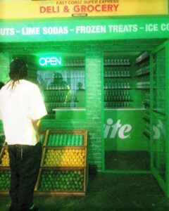 Sprite-casita-hollywood-brand-event - Bronson Hospitality Man with dreadlocks outside a green deli/grocery at night, next to a fruit display and neon OPEN sign; shelves of bottles inside the shop.