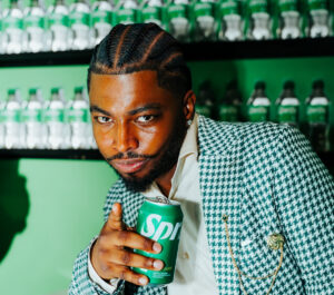 Sprite-casita-hollywood-brand-event - Bronson Hospitality Man with cornrowed hair and a patterned blazer holds a Sprite can in front of green shelves of Sprite bottles, looking at the camera.