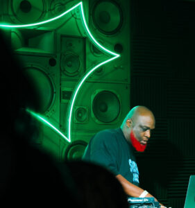 Sprite-casita-hollywood-brand-event - Bronson Hospitality DJ with a bright red beard at a booth, operating equipment in a room with a neon green sign and a wall of speakers behind him.