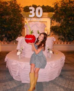 SnapInstato_684254100_17998567748930224_353721396570974838_n - Bronson Hospitality Woman in a black-and-white gingham dress posing with a giant red apple labeled 'philosophy' in front of a large pink cake prop.