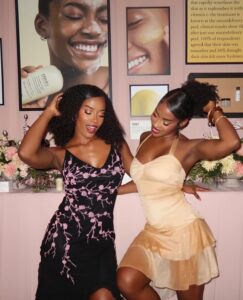 SnapInstato_684720463_17998567547930224_8951733674172263679_n - Bronson Hospitality Two women in party dresses dance together, smiling, in a pink-themed room with floral decor and framed wall images behind them.
