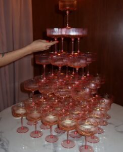 SnapInstato_684896651_17998567481930224_6879342106358520590_n - Bronson Hospitality Pink-tinted champagne coupe tower stacked on a marble table, with a hand placing the top glass on the pyramid.