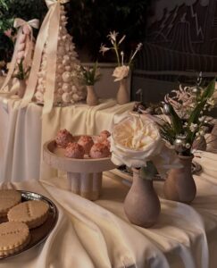 SnapInstato_686591974_17998567724930224_5740230766276938039_n - Bronson Hospitality Dessert table with pink frosted pastry balls on a pedestal, cream flowers in vases, and a plate of cookies on a white tablecloth