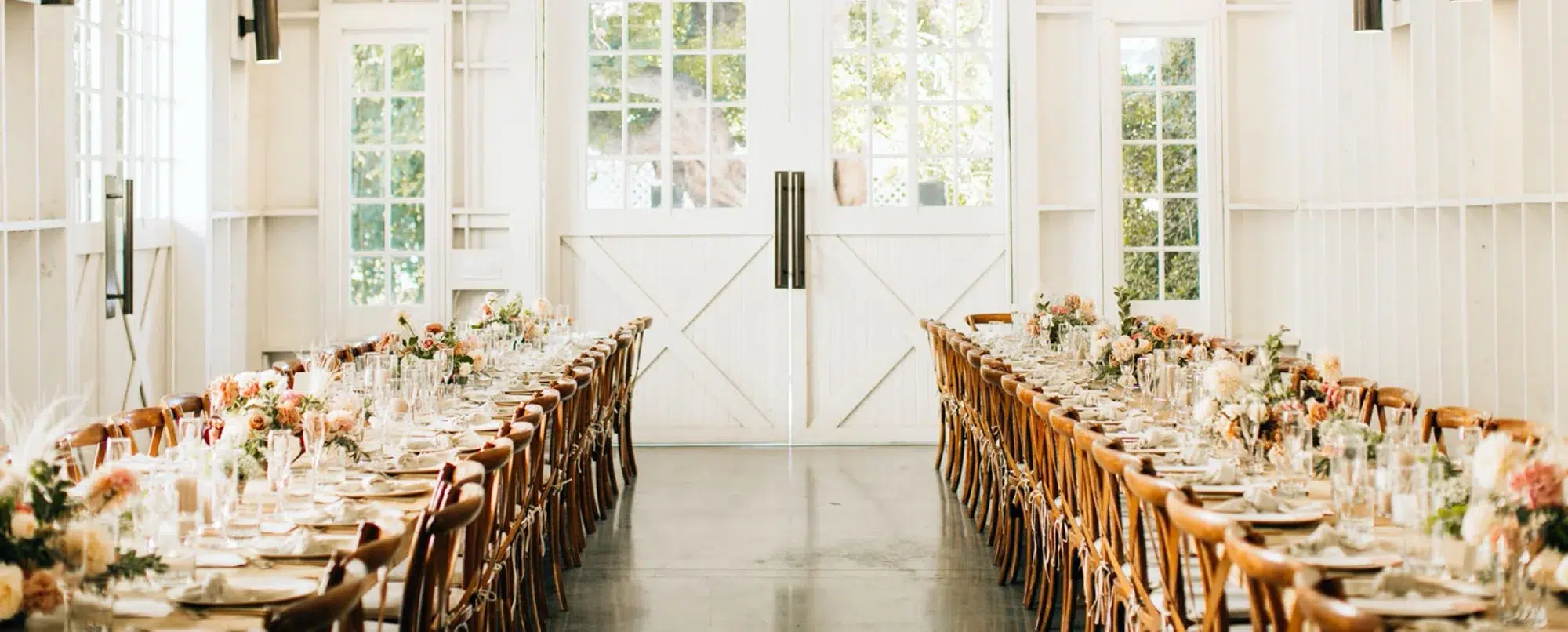 Bright white barn-style reception hall with long wooden tables and crossback chairs at Lombardi House, a Bronson Hospitality