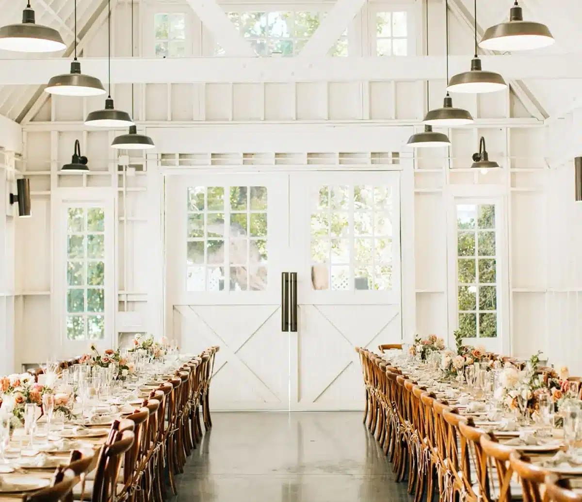 Bright white barn-style reception hall at Lombardi House with vaulted ceilings, pendant lighting, and long banquet tables set with floral centerpieces